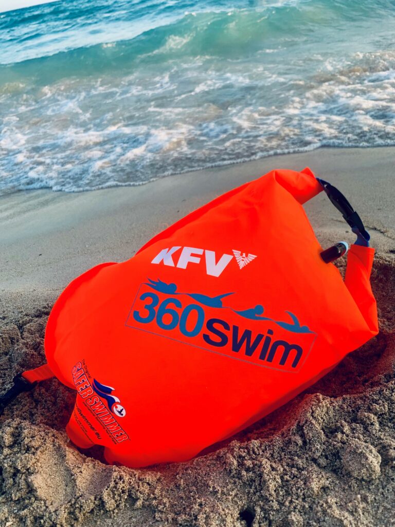Eine Schwimmboje im orangen Signalton liegt an einem Sandstrand. Der Aufdruck "KFV 360 swim" ist darauf zu sehen.