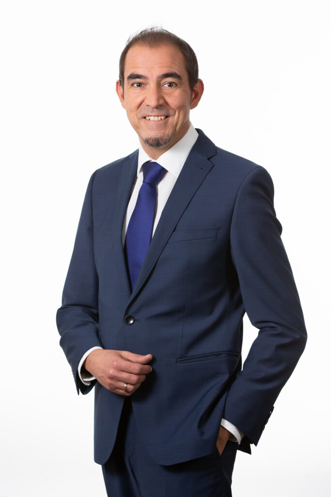 Ein Mann in dunklem Anzug mit weißem Hemd und dunkelblauer Krawatte.