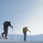 Zwei Skitourengehende auf einer beschneiten Piste mit Stöcken und Ausrüstung.