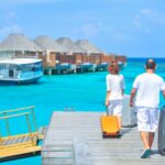 Zwei Urlauber*innen ziehen einen Koffer hinter sich über einen Steg her. Vor ihnen sieht man strahlend blaues Meer und Hütten sowie ein Boot.