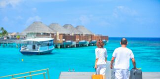 Zwei Urlauber*innen ziehen einen Koffer hinter sich über einen Steg her. Vor ihnen sieht man strahlend blaues Meer und Hütten sowie ein Boot.