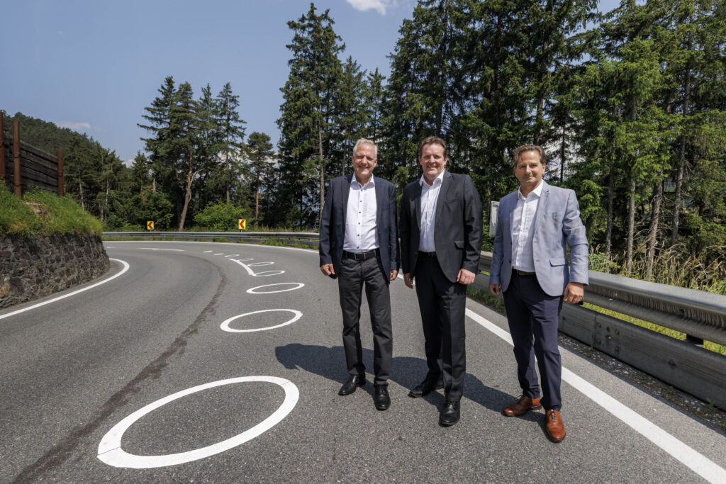 3 Männer in Anzügen stehen auf einer Straße in einer Kurve, die mit Ellipsen-Markierungen für Motorradfahrende gekennzeichnet ist.