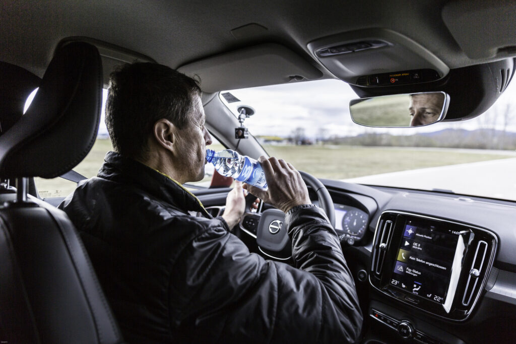 Ein Mann sitzt im Auto hinter einem Lenkrad und trinkt aus einer Wasserflasche.