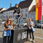 Zwei Mitarbeiter*innen bei der Verteilaktion, ausgestattet mit Taschen, neben einem Podium mit einem Auge und "nice to see you" lächeln in die Kamera. Auf dem Podium ist eine fahrradfahrende Puppe angebracht.