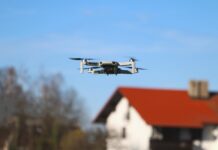 Eine Drohne fliegt im blauen Himmel. Im Hintergrund ist ein Haus und ein Baum verschwommen zu sehen.