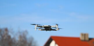 Eine Drohne fliegt im blauen Himmel. Im Hintergrund ist ein Haus und ein Baum verschwommen zu sehen.