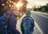 Zwei Buben auf dem Gehsteig neben einer Straße mit Schultasche, Jacke. Ein Bub blickt nach vorne Richtung Sonne, er trägt eine Haube. Ein Bub blickt nach hinten in die Kamera.