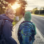 Zwei Buben auf dem Gehsteig neben einer Straße mit Schultasche, Jacke. Ein Bub blickt nach vorne Richtung Sonne, er trägt eine Haube. Ein Bub blickt nach hinten in die Kamera.