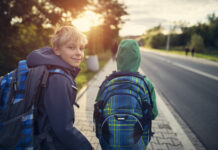 Zwei Buben auf dem Gehsteig neben einer Straße mit Schultasche, Jacke. Ein Bub blickt nach vorne Richtung Sonne, er trägt eine Haube. Ein Bub blickt nach hinten in die Kamera.