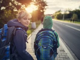 Zwei Buben auf dem Gehsteig neben einer Straße mit Schultasche, Jacke. Ein Bub blickt nach vorne Richtung Sonne, er trägt eine Haube. Ein Bub blickt nach hinten in die Kamera.