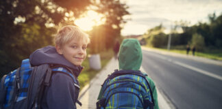 Zwei Buben auf dem Gehsteig neben einer Straße mit Schultasche, Jacke. Ein Bub blickt nach vorne Richtung Sonne, er trägt eine Haube. Ein Bub blickt nach hinten in die Kamera.