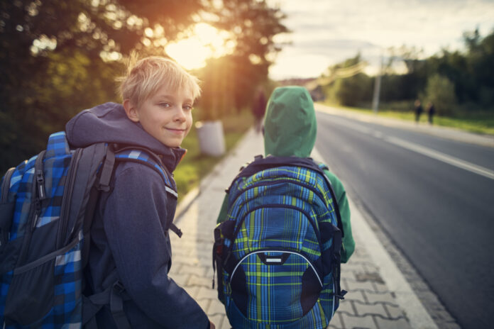 Zwei Buben auf dem Gehsteig neben einer Straße mit Schultasche, Jacke. Ein Bub blickt nach vorne Richtung Sonne, er trägt eine Haube. Ein Bub blickt nach hinten in die Kamera.