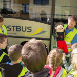 Kinder stehen vor dem Bus bei der Aktion. Ein Mann zeigt Sicherheitsmaßnahmen mit einer Handpuppe vor.