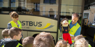 Kinder stehen vor dem Bus bei der Aktion. Ein Mann zeigt Sicherheitsmaßnahmen mit einer Handpuppe vor.