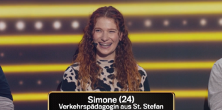 KFV-Mitarbeiterin Simone bei der Quiz-Show. Sie steht an einem Podium und lacht herzlich. Sie hat lange, lockige, rötliche Haare und trägt schwarz-weiß gemusterte Kleidung. Sie trägt ein Mikrofon an ihrer rechten Wange bzw. hinter ihrem rechten Ohr. Im Hintergrund sind gelbe Linien und Lichter zu sehen. Im Bild eingeblendet ist der Text: "Simone (24) Verkehrspädagogin aus St. Stefan"