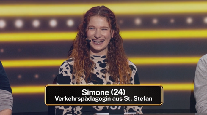 KFV-Mitarbeiterin Simone bei der Quiz-Show. Sie steht an einem Podium und lacht herzlich. Sie hat lange, lockige, rötliche Haare und trägt schwarz-weiß gemusterte Kleidung. Sie trägt ein Mikrofon an ihrer rechten Wange bzw. hinter ihrem rechten Ohr. Im Hintergrund sind gelbe Linien und Lichter zu sehen. Im Bild eingeblendet ist der Text: 