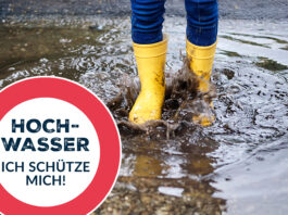 Die Füße einer Person in gelben Gummistiefeln ist zu sehen, die durch Wasser watet. Daneben das Warnschild "Hochwasser - ich schütze mich"