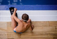 Ein Junge (ca.10-12 Jahre) mit Taucherflossen und vermutlich Schwimmbrille sitzt am Beckenrand. Das Bild ist in Vogelperspektive aufgenommen.
