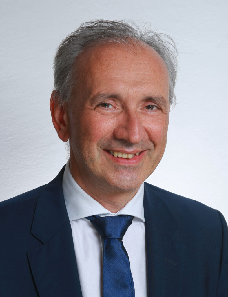Ein weißer Mann mittleren Alters in Anzug und Kravatte lächelt in die Kamera.