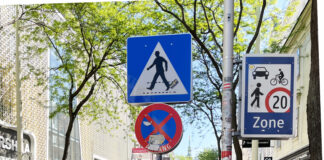 Eine Begegnungszone: Sichtbar sind die Passant*innen und die Schilder für die zu Fuß Gehenden und die begrünte Begegnungszone.