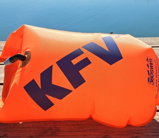Eine orange Schwimmboje mit KFV-Logo liegt aufgeblasen auf dem Holzsteg vor einem spiegelglatten See.