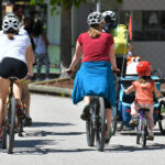 Man sieht Radfahrer*innen von hinten: Kinder wie erwachsene, selbst- sowie mitfahrend, auf einer Straße.