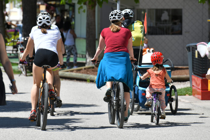 Man sieht Radfahrer*innen von hinten: Kinder wie erwachsene, selbst- sowie mitfahrend, auf einer Straße.