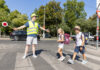 Ein Schülerlotse mit Warnweste und gelber Kappe hilft 2 Schulkindern über den Zebrastreifen. Er hält dazu auch einen Signalstab.