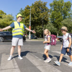 Ein Schülerlotse mit Warnweste und gelber Kappe hilft 2 Schulkindern über den Zebrastreifen. Er hält dazu auch einen Signalstab.