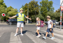 Ein Schülerlotse mit Warnweste und gelber Kappe hilft 2 Schulkindern über den Zebrastreifen. Er hält dazu auch einen Signalstab.