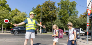 Ein Schülerlotse mit Warnweste und gelber Kappe hilft 2 Schulkindern über den Zebrastreifen. Er hält dazu auch einen Signalstab.