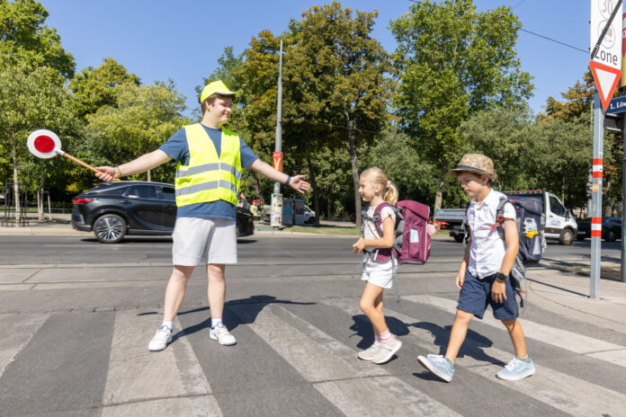 Ein Schülerlotse mit Warnweste und gelber Kappe hilft 2 Schulkindern über den Zebrastreifen. Er hält dazu auch einen Signalstab.