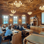 Teilnehmende einer Tagung auf mehreren Bänken in einem Vortragssaal sitzen. Der Saal ist prunkvoll mit schönen Lustern und einer kunstvollen Decken-Verzierung.