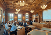 Teilnehmende einer Tagung auf mehreren Bänken in einem Vortragssaal sitzen. Der Saal ist prunkvoll mit schönen Lustern und einer kunstvollen Decken-Verzierung.