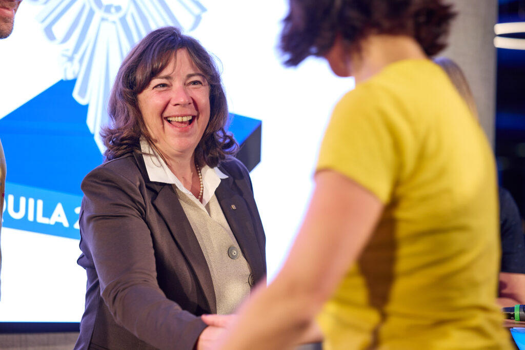 Eine Frau im Hosenanzug und Hemd schüttelt freudestrahlend einer Person in gelbem T-Shirt die Hand.