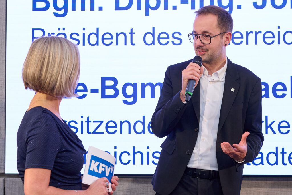 Ein Mann in Anzug und Hemd sowie Brille spricht zu einer blonden Frau mit Moderationskarten in ein Mikrofon.