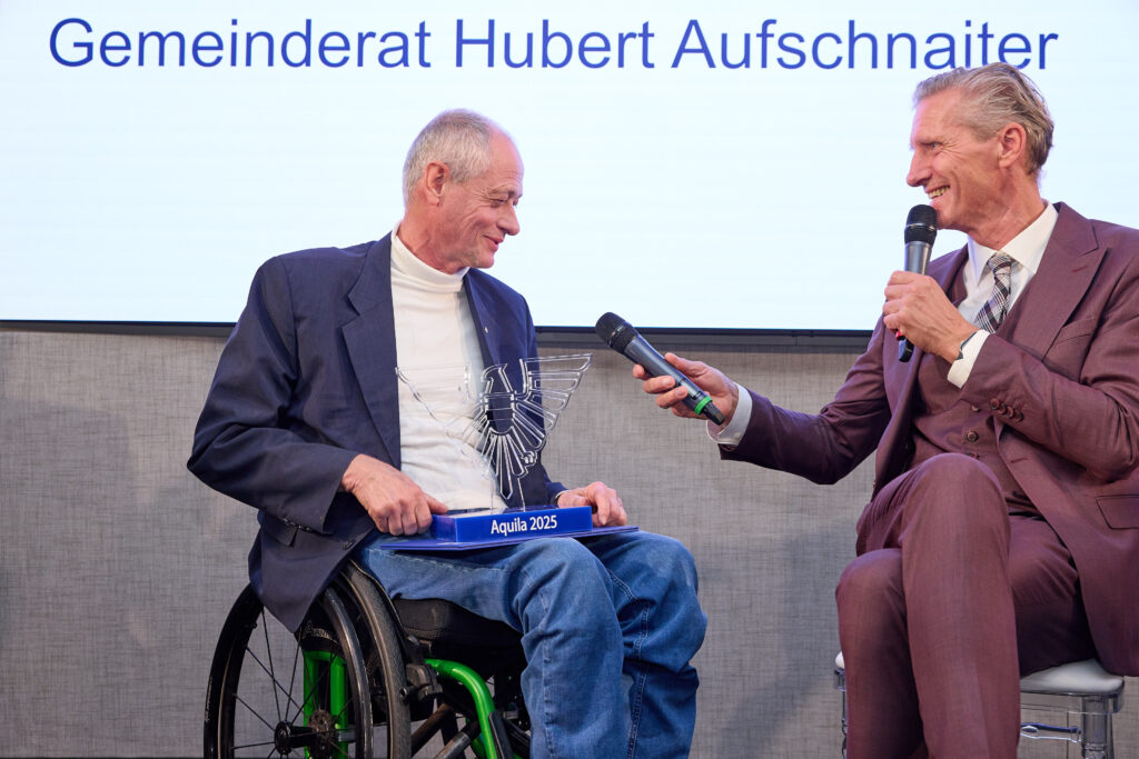 in Mann im Rollkragenpulli und Jackett sitzt im Rollstuhl und hat den Preis "Aquila 2025" auf seinem Schoß abgestellt. Moderator Oliver Zeisberger sitzt neben ihm auf einem Sessel und hält ihm lachend ein Mikrofon hin.