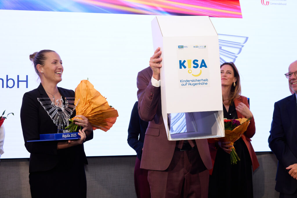 Eine Frau mit AQUILA-Trophäe und Blumenstrauß strahlt und blickt zu einem Mann, der ein weißes Periscope vor den Körper hält, auf dem "K!SA" aufgedruckt steht.