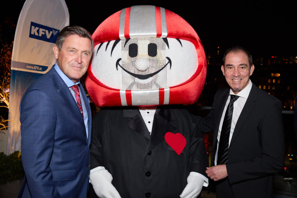 Ein HELMI-Maskottchen im schwarzen Anzug zwischen Mobilitätsminister Peter Hanke und KFV-Direktor Christian Schimanofsky, ebenfalls im Anzug, lächeln in die Kamera.