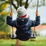 Zwei Drittel aller Kinderunfälle sind Stürze – Herbst besonders unfallreich Man sieht ein Kind von hinten, das auf einer Schaukel sitzt. Das Kind trägt eine graue Haube und eine dunkelblaue, gefütterte Jacke. Auch die Stimmung im Hintergrund deutet auf Herbst hin, mit grauem Himmel und teilweise verfärbtem Laub.
