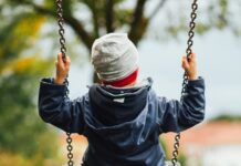Man sieht ein Kind von hinten, das auf einer Schaukel sitzt. Das Kind trägt eine graue Haube und eine dunkelblaue, gefütterte Jacke. Auch die Stimmung im Hintergrund deutet auf Herbst hin, mit grauem Himmel und teilweise verfärbtem Laub.