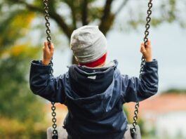 Man sieht ein Kind von hinten, das auf einer Schaukel sitzt. Das Kind trägt eine graue Haube und eine dunkelblaue, gefütterte Jacke. Auch die Stimmung im Hintergrund deutet auf Herbst hin, mit grauem Himmel und teilweise verfärbtem Laub.