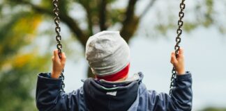 Man sieht ein Kind von hinten, das auf einer Schaukel sitzt. Das Kind trägt eine graue Haube und eine dunkelblaue, gefütterte Jacke. Auch die Stimmung im Hintergrund deutet auf Herbst hin, mit grauem Himmel und teilweise verfärbtem Laub.