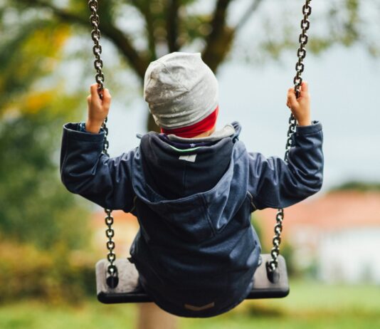 Man sieht ein Kind von hinten, das auf einer Schaukel sitzt. Das Kind trägt eine graue Haube und eine dunkelblaue, gefütterte Jacke. Auch die Stimmung im Hintergrund deutet auf Herbst hin, mit grauem Himmel und teilweise verfärbtem Laub.