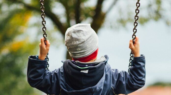 Zwei Drittel aller Kinderunfälle sind Stürze – Herbst besonders unfallreich Man sieht ein Kind von hinten, das auf einer Schaukel sitzt. Das Kind trägt eine graue Haube und eine dunkelblaue, gefütterte Jacke. Auch die Stimmung im Hintergrund deutet auf Herbst hin, mit grauem Himmel und teilweise verfärbtem Laub.