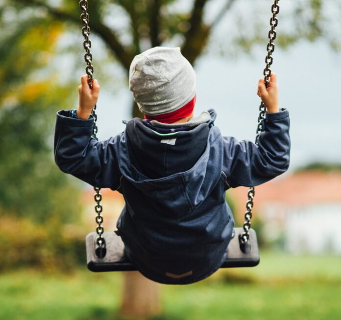 Man sieht ein Kind von hinten, das auf einer Schaukel sitzt. Das Kind trägt eine graue Haube und eine dunkelblaue, gefütterte Jacke. Auch die Stimmung im Hintergrund deutet auf Herbst hin, mit grauem Himmel und teilweise verfärbtem Laub.