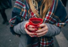 Gefährliche Adventwochen: Mehr Unfälle durch Alkohol im Straßenverkehr Eine Person mit langen blonden Haaren in Wintermantel mit einem karierten Schal hält ein herzförmiges Punsch-Häferl in Händen mit rot manikürten Nägeln. Fokus ist auf das Punschhäferl, Gesicht und Beine sind nicht zu sehen. Im Hintergrund sind die Beine anderer Leute zu sehen, die auf Asphalt stehen.