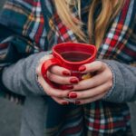 Eine Person mit langen blonden Haaren in Wintermantel mit einem karierten Schal hält ein herzförmiges Punsch-Häferl in Händen mit rot manikürten Nägeln. Fokus ist auf das Punschhäferl, Gesicht und Beine sind nicht zu sehen. Im Hintergrund sind die Beine anderer Leute zu sehen, die auf Asphalt stehen.