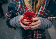 Eine Person mit langen blonden Haaren in Wintermantel mit einem karierten Schal hält ein herzförmiges Punsch-Häferl in Händen mit rot manikürten Nägeln. Fokus ist auf das Punschhäferl, Gesicht und Beine sind nicht zu sehen. Im Hintergrund sind die Beine anderer Leute zu sehen, die auf Asphalt stehen.