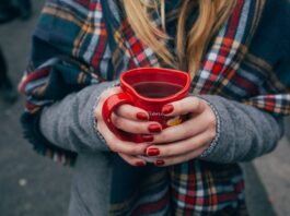 Eine Person mit langen blonden Haaren in Wintermantel mit einem karierten Schal hält ein herzförmiges Punsch-Häferl in Händen mit rot manikürten Nägeln. Fokus ist auf das Punschhäferl, Gesicht und Beine sind nicht zu sehen. Im Hintergrund sind die Beine anderer Leute zu sehen, die auf Asphalt stehen.